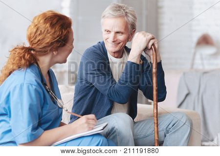 Possitive attitude to patients. Relaxed retired man with a walking stick smiling while sitting on a sofa and talking to a female doctor during a regular visit at home.