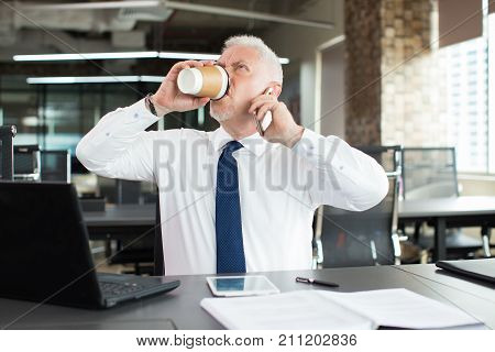 Crazy businessman drinking coffee avidly at work to have more energy. Angry tired senior manager talking on phone. Overworked concept