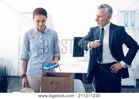 Doors are there. Angry serious businessman standing next to his fired young employee packing her stuff in a box and pointing at a door while waiting for her to leave the office.