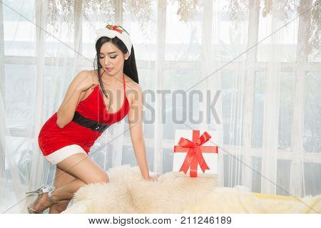Pretty Asian woman in Santa dresses with gift kneel on the sofa in a room.