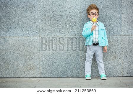 Fashion baby boy in mint jacket and sneakers stands on a gray wall background. Trendy boy with lollipop standing on the street.