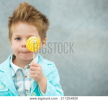 Fashion baby boy in mint jacket and sneakers stands on a gray wall background. Trendy boy with lollipop standing on the street.