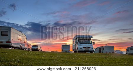 Caravans and cars parked on a grassy campground in summer under beautiful sunset