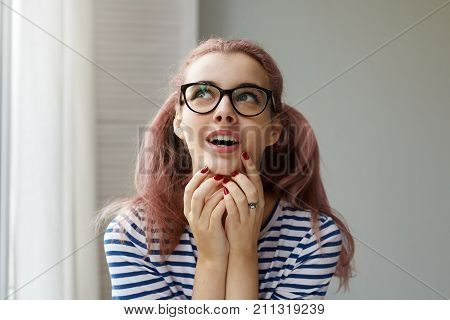 Headshot of smart cunning young woman in black rimmed glasses and striped top touching her face and looking up with sly or mysterious expression opening mouth while planning or scheming something