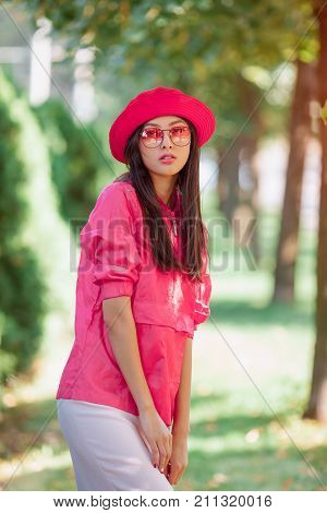 Asian woman fashion portrait. Beautiful mixed race asian caucasian young girl in pink hat jaket and red sunglasses looking at camera outdoor against green blurred bokeh nature background. Gorgeous slim model in trendy casual colourful clothes posing outsi