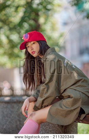 Asian woman urban fashion photo. Beautiful mixed race asian caucasian young girl in red cap green raincoat looking at camera outdoor against green blurred bokeh city background. Gorgeous slim model in trendy casual colourful clothes sitting on bench outsi