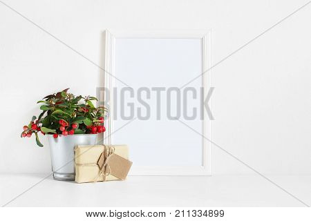 White blank wooden frame mockup with potted red teaberry plant and package with a gift tag on the white table. Styled stock feminine photography. Home decor, Christmas winter concept.