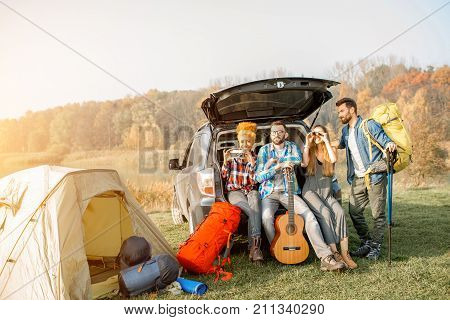 Multi ethnic group of friends sitting at the car trank during the outdoor recreation with tent and hiking equipment near the forest