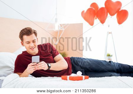 Full of love. Cheerful young man smiling while relaxing on his bed and messaging with his girlfriend on Valentines Day.