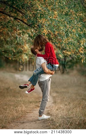 Young man lifted up girl in his hands and they stand under tree and kiss on walk in forest.