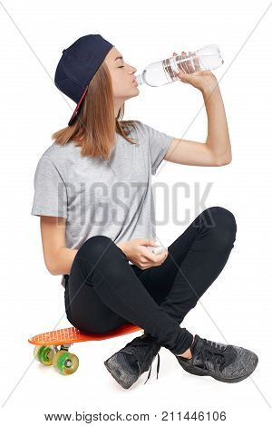 Teen girl in full length sitting on skate board drinking water from a bottle enjoying with closed eyes isolated on white background