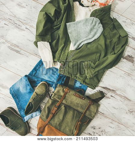 Flat-lay of Fall lady' s clothes outfit. Ecru colored knitted woolen sweater, parka, linen scarf, denim jeans, haki suede boots, backpack, cap over parquet background, top view, square crop