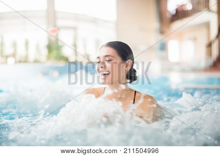 Excited female laughing while splashing warm water during spa procedure in whirlpool