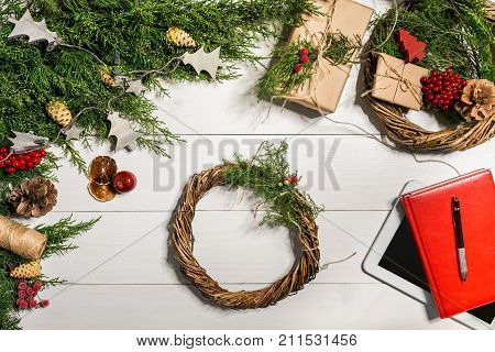 Juniper branches with a Christmas decor. Christmas, New Year background. Coniferous branches of juniper, red notepad and white tablet with black screen. Top view, flat design. Christmas tree on white wooden background.