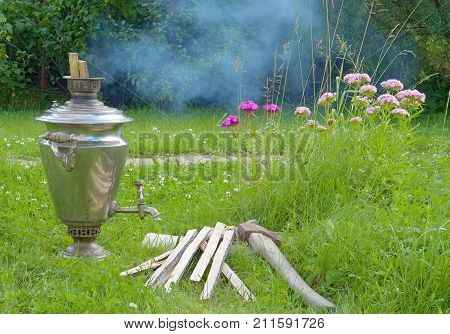 shiny samovar is heated by the fire burning splinters of wood and it emits white smoke; and the samovar standing on a green grass on a background of different colors of red and pink next to a samovar lies the axe and the wood. day in Russia closeup
