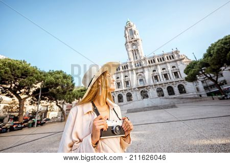 Portrait of a young woman tourist in sunhat standing with photo camera in front of the city hall building during the morning light in Porto, Portugal