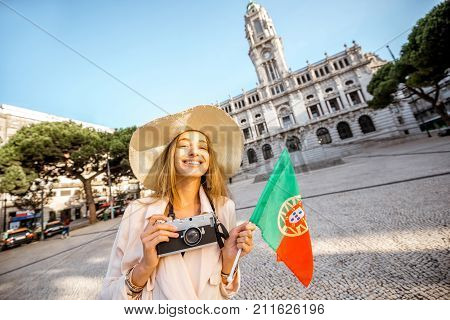 Portrait of a young woman tourist in sunhat standing with portuguese flag in front of the city hall building during the morning light in Porto, Portugal