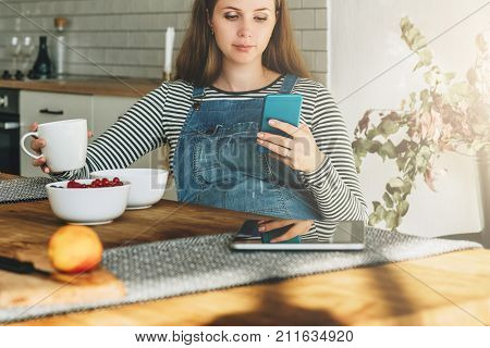A young pregnant woman is sitting in the kitchen at the table, having breakfast, drinking tea and using a smartphone. Lifestyle, social network. Online marketing, education, blogging, chatting.