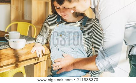 View from above.Morning, breakfast.Young married couple in kitchen.Pregnant woman is sitting at table, man is holding her pregnant belly.On table is laptop, tablet computer, dishes. Lifestyle.