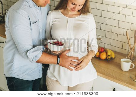 Young married couple embraces standing near table in kitchen. Husband hugs his pregnant wife, putting his hands on her big belly. A loving couple, future parents. Lifestyle, happy people.