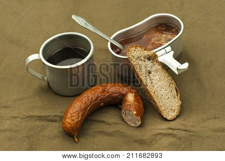 a scanty road meal: soup in a mess tin coffee in a vintage army mug a loaf of bread and a piece of sausage on a spread tarpaulin
