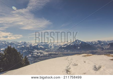 Scenery of footprint in snow on cliff with rock mountain range in winter background under blue clear sky and wonderful cloud shape, Landscape of Swiss Alps hill in Switzerland behind snowy ground stamp by animal spoor