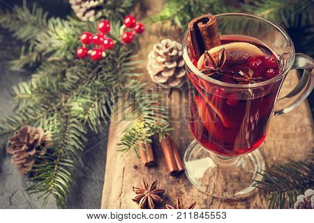 Hot mulled wine drink with lemon, apple, cinnamon, anise and other spices in a glass cup between fir tree branches on wooden cutting board