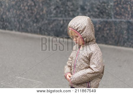 Little Cute  Caucasian Blonde Girl In A Hooded Jacket Upset Or Offended Standing Alone On A City Str