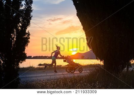 Running mother with child in stroller enjoying motherhood at sunset and mountains landscape. Jogging or power walking woman with pram on a beach at sunset. Beautiful inspirational mountains landscape.