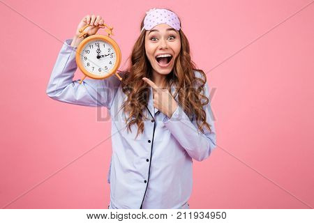 Portrait of an excited young girl dressed in pajamas pointing at alarm clock and looking at camera isolated over pink background