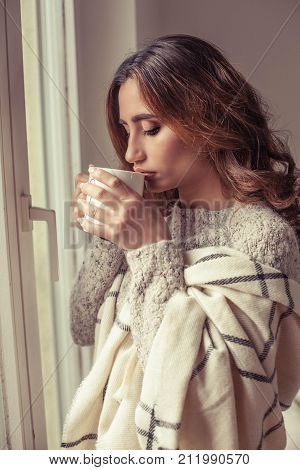 Young Beautiful Woman Stands Near A Window, Wrapped In A Blanket And Drink Hot Coffee. Cozy. Autumn.