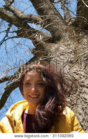 Close Up Outdoos Portrait Of Young Smilling Girl In Yellow Jacket Sitting Near Big Old Tree In The P