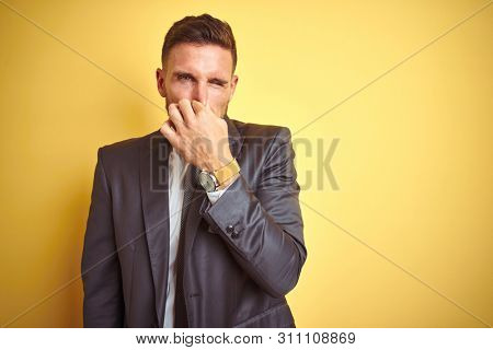 Young handsome business man over yellow isolated background smelling something stinky and disgusting, intolerable smell, holding breath with fingers on nose. Bad smells concept.