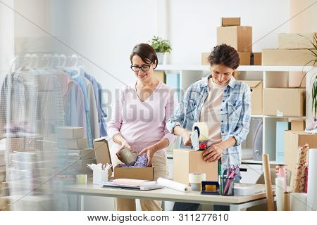 Two Young Women Packing Parcels For Online Clients - Putting Things In Boxes And Sealing Them With C