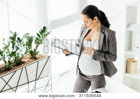High Angle View Of Pregnant Woman Looking At Folder While Standing In Office Near Flowerpot With Pla