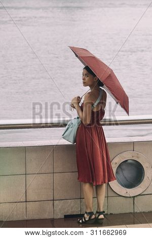 Asian woman walking by river with umbrella under rain during rainy season on summer day vacation. Elegant chinese lady in red dress.
