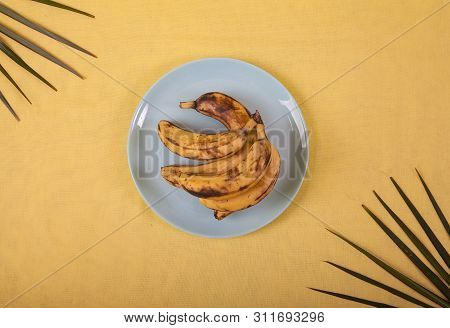 Ugly Bananas Lie On A Blue Porcelain Plate On A Yellow Background With Palm Leaves