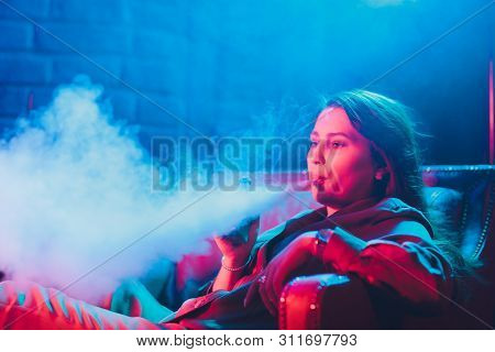 Woman Smokes A E-cigarette Lying On The Bed In The Room. Smokers Concept Vaping.