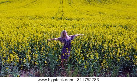 Happy Blonde Girl In Dress With Flower Print On The Blooming Yellow Rapeseed Field