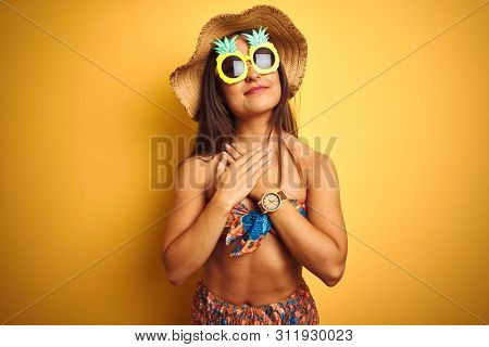 Woman on vacation wearing bikini and pineapple sunglasses over isolated yellow background smiling with hands on chest with closed eyes and grateful gesture on face. Health concept.