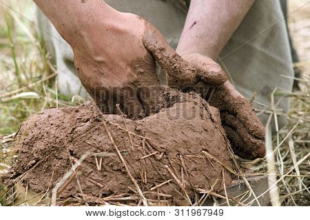 The Hands Of A Man In Historical Clothes Making A Furnace Of Clay And Straw In The Open Water. Festi
