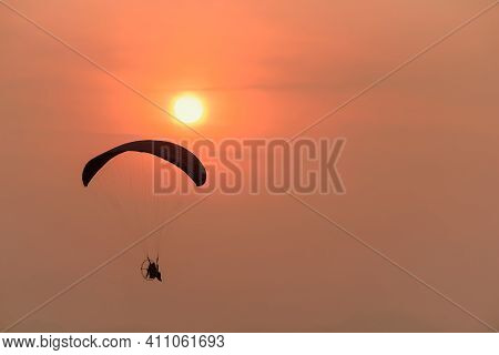 Silhouette Of The Paramotor Gliding And Flying In The Air Through Soft Sunlight Sunset Sky.