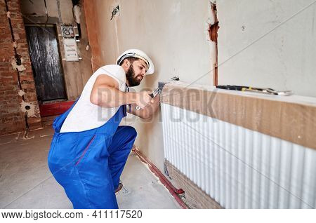 Young Plumber, Wearing Blue Overalls, White Helmet, Installing Water Radiator, Working With Wrench. 
