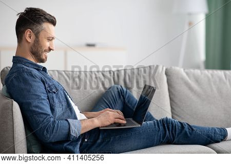 Side View Of Middle-aged Man Reclining On Couch In Living Room, Using Newest Laptop, Copy Space. Hap
