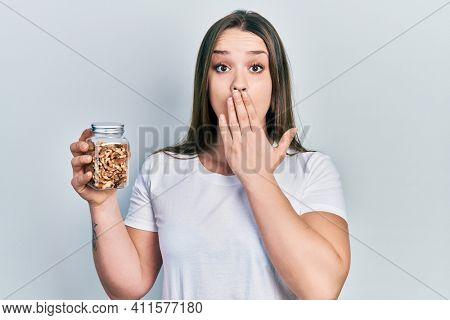 Young hispanic girl holding jar with walnuts covering mouth with hand, shocked and afraid for mistake. surprised expression 
