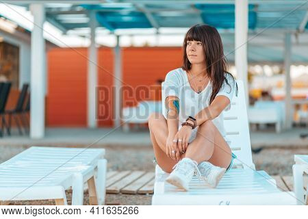 Tanned Pretty Woman With Tattoos, Posing Sitting On A Chaise Longue. Waterfront Cafes And The Beach.