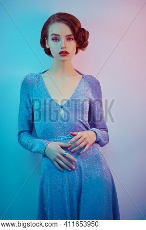Beautiful melancholic girl in an elegant dress stands against mixed blue background. Hair and makeup in the style of the 20s. 