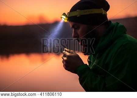 Side View Of Bearded Male Traveler In Warm Clothing And Glowing Headlamp Warming Up Hands With Breat
