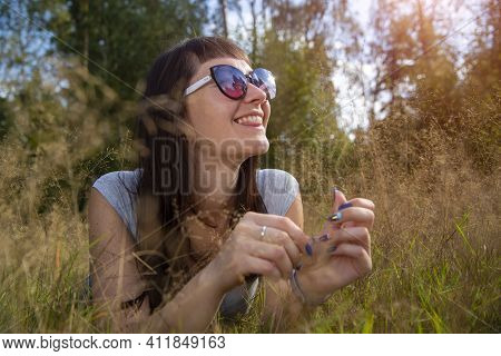 Happy Beautiful Young Woman In Sunglasses Smiles And Lies On A Meadow In The Grass In The Sunshine E
