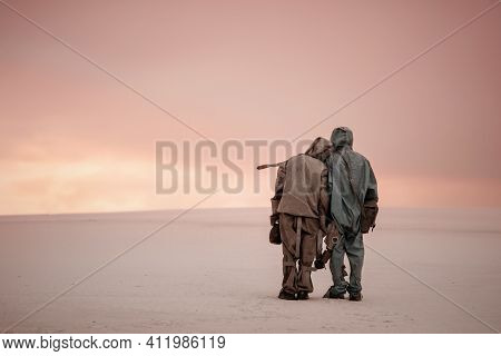 A Man And A Woman In Chemical Protection Suits Stand Side By Side On A Sandy Landscape. In The Hands
